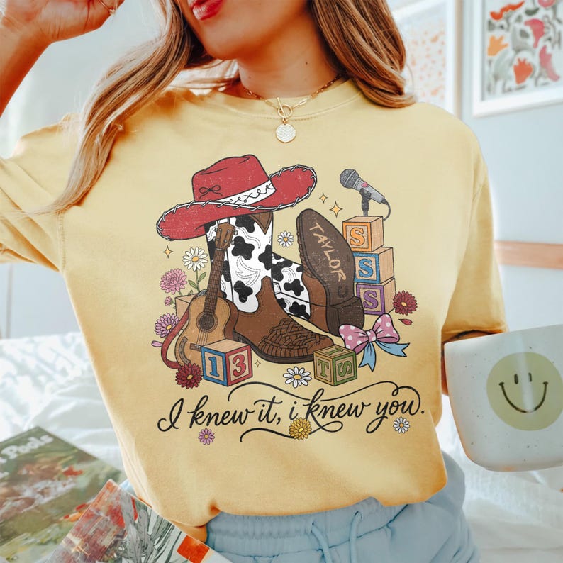 Pu&ograve; includere: Una t-shirt girocollo giallo pallido con una stampa grafica raffigurante un cappello da cowboy rosso, stivali con stampa mucca, una chitarra acustica, un microfono, cubi dell'alfabeto e un fiocco. Il testo recita "TAYLOR" e "I knew it, i knew you."