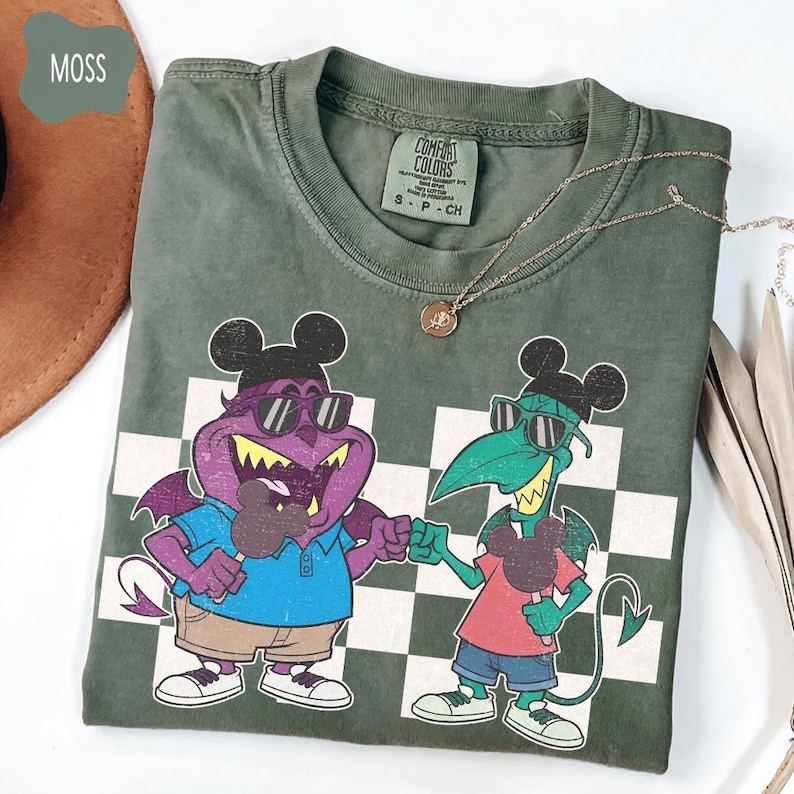 May include: Moss green Comfort Colors t-shirt featuring a cartoon graphic of two devilish characters with Mickey Mouse ears, sunglasses, and ice cream. The shirt has a checkered pattern and a gold necklace.