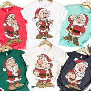 Op de afbeelding: Een verzameling van zes shirts met korte mouwen in verschillende kleuren, elk met een cartoon kerstman karakter omwikkeld met kerstverlichting. De shirts zijn rood, wit, groen, lichtblauw en antracietgrijs.