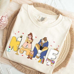 Peut inclure: Un t-shirt couleur crème arborant le mot NAME en grandes lettres style bloc. Les lettres sont décorées d'illustrations de personnages de La Belle et la Bête, incluant Belle, la Bête, Lumière, Big Ben et Madame Samovar.