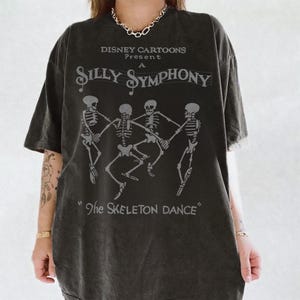 May include: A charcoal grey short-sleeve t-shirt featuring a graphic print of four skeletons dancing. The text reads "DISNEY CARTOONS Present A SILLY SYMPHONY" and "The SKELETON DANCE" in a vintage-style font. The shirt has a crew neckline.