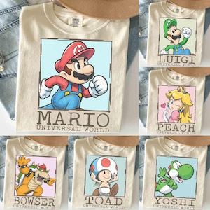 May include: Cream-colored t-shirts featuring various characters from the Mario video game series. Each shirt displays a character's image within a square frame, with the character's name and "UNIVERSAL WORLD" printed below. Characters include Mario, Luigi, Peach, Bowser, Toad, and Yoshi.