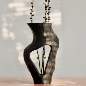 May include: A black, textured vase with a unique, open design. The vase has a geometric pattern and holds delicate, green-stemmed branches with small, round buds. The vase is a decorative item, perfect for displaying floral arrangements or as a standalone art piece.