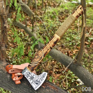 May include: Axe with a wooden handle wrapped in twine. The axe head is silver with etched designs and a leather sheath. The handle has carved details. The axe is resting on a tree branch.