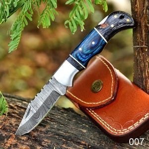 May include: A Damascus steel knife with a blue and black handle and a brown leather sheath. The knife has a serrated edge and a silver bolster. The handle is decorated with white and gold accents. The sheath is closed with a brass button.