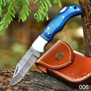 Peut inclure: Un couteau pliant avec un manche en bois bleu et une lame à motifs, posé sur une surface en bois. Le couteau a une mitre argentée et est associé à un étui en cuir marron. Le numéro "006" est visible.