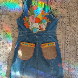 May include: A blue denim dress with a fruit basket embroidered on the back. The dress has two pockets with brown fabric and fruit embroidery on the top.