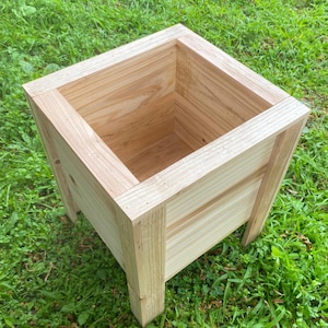 May include: A square wooden planter box made from light-coloured wood. The planter has a simple, open design with four legs. The box is empty and ready for plants. The planter is sitting on green grass.