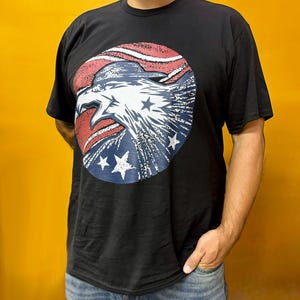 May include: Black t-shirt featuring a circular graphic of an eagle's head with red and white stripes and blue stars. The eagle's head is white with detailed feathering. The shirt is a classic crew neck style.