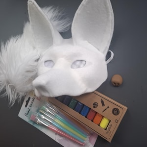 Può includere: Una maschera da volpe bianca con grandi orecchie, un piccolo naso marrone e un ciuffo di piume bianche. Il kit include pennelli, una tavolozza di colori e una scatola fai-da-te con il testo "Acrylfarbe".