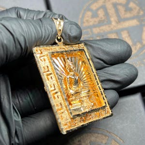 May include: A gold-colored pendant featuring a seated Buddha figure within a rectangular frame. The pendant has intricate detailing and a textured surface. The Buddha is in a meditative pose, with a radiant halo behind it. The pendant is suspended from a matching gold bail.