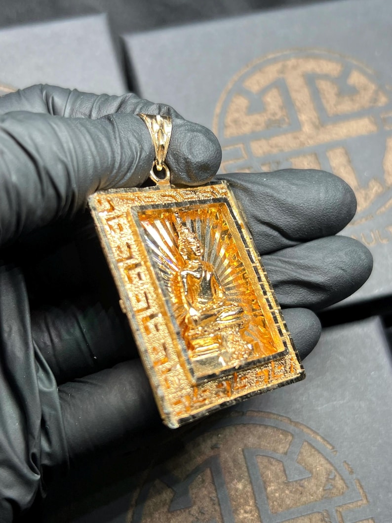 May include: A gold-colored pendant featuring a seated Buddha figure within a rectangular frame. The pendant has intricate detailing and a textured surface. The Buddha is in a meditative pose, with a radiant halo behind it. The pendant is suspended from a matching gold bail.