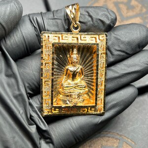 May include: A gold-colored pendant featuring a seated Buddha figure within a rectangular frame. The frame has an ornate, geometric border. The Buddha is in a meditative pose, with a radiant background. The pendant has a loop for a chain.