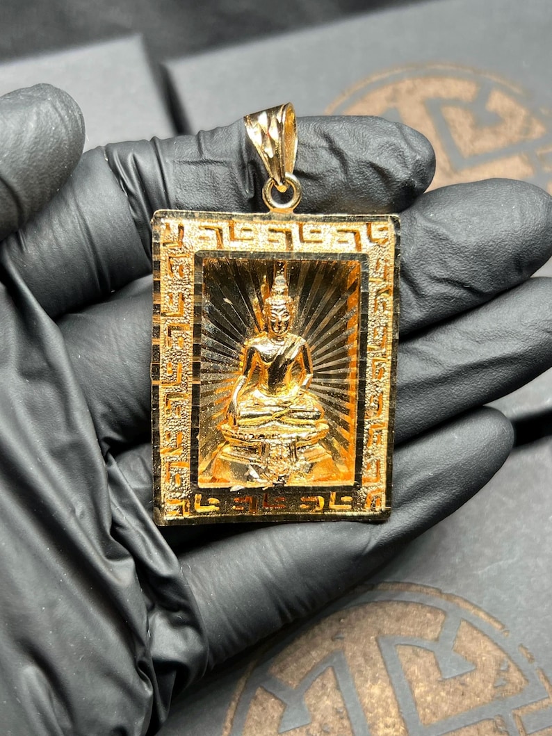 May include: A gold-colored pendant featuring a seated Buddha figure within a rectangular frame. The frame has an ornate, geometric border. The Buddha is in a meditative pose, with a radiant background. The pendant has a loop for a chain.