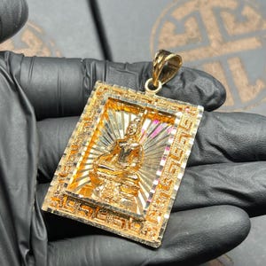 May include: A gold-colored pendant featuring a detailed Buddha figure seated in a meditative pose. The rectangular pendant has an ornate frame with intricate patterns and a bail for a chain. The background has a sunburst design.