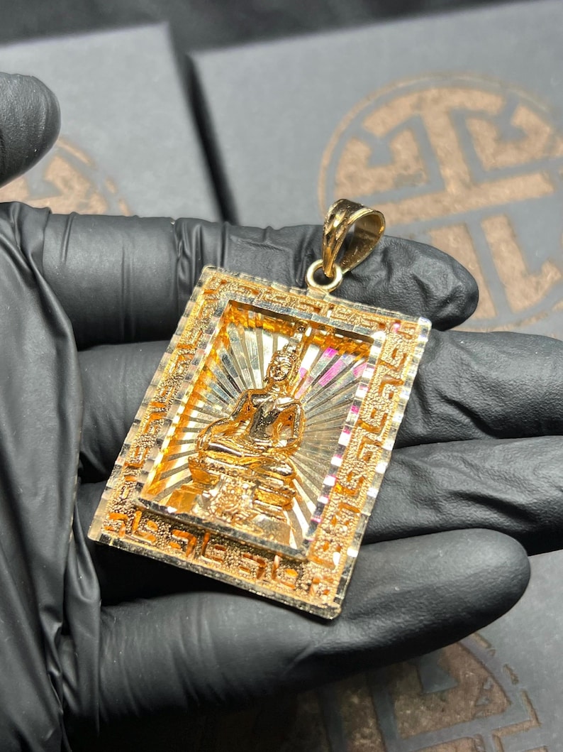 May include: A gold-colored pendant featuring a detailed Buddha figure seated in a meditative pose. The rectangular pendant has an ornate frame with intricate patterns and a bail for a chain. The background has a sunburst design.