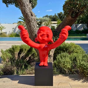 Peut inclure: Une sculpture rouge vif représentant une figurine souriante aux bras tendus, montée sur un cube noir. L'œuvre est située à l'extérieur, entourée de verdure et d'un ciel bleu, créant un contraste saisissant.