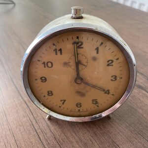May include: A vintage silver-toned alarm clock with a cream-colored face. The clock features black numerals, a small seconds dial, and two silver-toned legs. The hands point to approximately 4:00. The clock has a silver-toned top button.