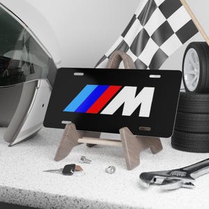 May include: Black license plate with a white "M" and a blue, red, and blue logo. The plate is on a wooden stand.