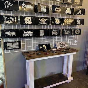 May include: A display of black license plates with various designs and text, including "BEEF", "Braves", and "TOYODA". The plates are mounted on a wire grid, with a wooden table below displaying smaller items.