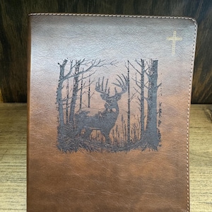 May include: Brown leather-bound book with a detailed forest scene featuring a deer. The cover has a gold cross in the upper right corner. The book's edges are stitched with white thread.