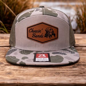 May include: A flat bill snapback hat with a camouflage pattern in shades of green and gray. The hat features a brown leather patch with the words "Chasin' Bands" and a dog graphic. The hat has a mesh back and a flat bill.
