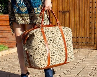 Puede incluir: Una bolsa de viaje beige y negra estampada con detalles y asas de cuero marrón. La bolsa tiene forma rectangular y cierre de cremallera. La bolsa es sostenida por una persona que lleva un vestido azul estampado y tacones abiertos azules.