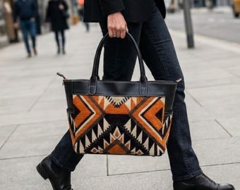May include: A large tote bag featuring a black leather top section and handles, with a lower body displaying a geometric pattern in shades of orange, black, white, and tan. The bag has two side pockets and visible stitching along the leather trim.