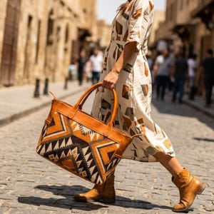 May include: A large tote bag featuring a tan leather top and handles with a woven fabric body displaying a geometric pattern in shades of brown, black, and cream. The bag is paired with a cream-colored dress with brown shapes and tan leather boots.