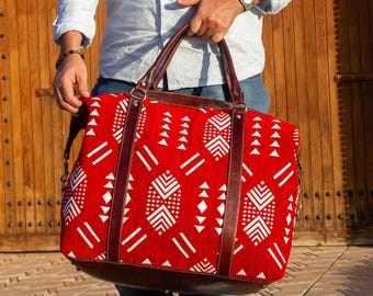 May include: A red and white patterned tote bag with brown leather handles and trim. The bag has a geometric design and a brown leather base. The bag is being held by a person wearing a light blue shirt and jeans.