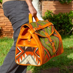 Könnte beinhalten: Ein orangefarbener Rucksack mit einem geometrischen Muster in Grün, Orange und Grau. Er hat braune Lederakzente, einen oberen Griff und verstellbare Riemen. Der Rucksack ist ideal für den Transport von Gegenständen.