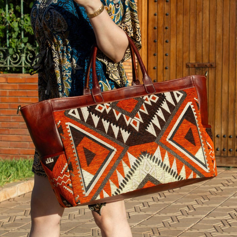 May include: A large tote bag featuring brown leather handles and side panels, combined with a central fabric panel displaying a geometric pattern in shades of burnt orange, dark brown, and cream, with two leather top handles and side pockets.