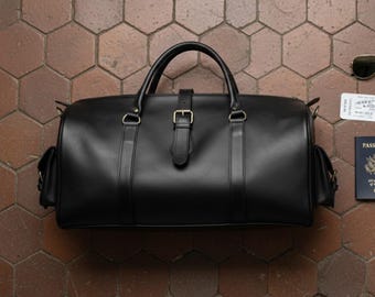 Può includere: Una borsa da viaggio in pelle nera con manico superiore e tracolla. La borsa ha una chiusura con fibbia e due tasche laterali. Occhiali da sole, una carta d'imbarco e un passaporto sono accanto alla borsa.