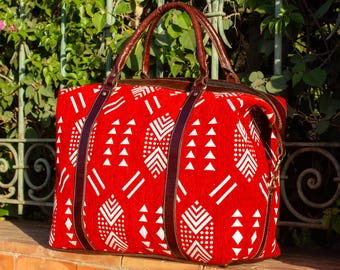 May include: A red travel bag with a white geometric pattern. The bag has brown leather handles and straps. The bag is sitting on a brick wall in front of a green leafy background.
