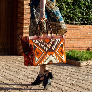 May include: A large rectangular tote bag featuring a central panel with a geometric pattern in orange, brown, white, and black. The bag has dark brown leather handles and trim, with a side pocket displaying a matching red and black patterned fabric.