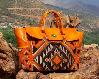 May include: A structured handbag featuring smooth orange leather handles and top trim, combined with a central panel of woven fabric displaying a geometric pattern in shades of orange, dark brown, and cream, secured with a gold-tone metal turn-lock.