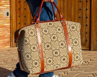 May include: A beige and black patterned travel bag with brown leather handles and accents. The bag is rectangular with a zipper closure and is being carried by a person wearing blue jeans and white sneakers.