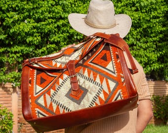May include: A large, rectangular travel bag with a geometric pattern in orange, white, and gray. The bag has brown leather accents, straps, and a buckle closure. A beige cowboy hat is visible above the bag.