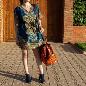 May include: A short-sleeved tunic dress featuring a deep V-neckline and a drawstring waist, decorated with a blue, gold, and black paisley pattern. A large brown leather shoulder bag with a woven orange and black geometric base is carried.