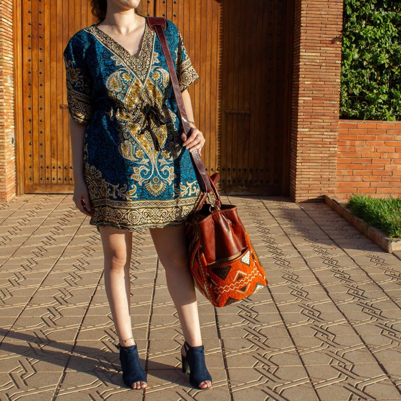 May include: A short-sleeved tunic dress featuring a deep V-neckline and a drawstring waist, decorated with a blue, gold, and black paisley pattern. A large brown leather shoulder bag with a woven orange and black geometric base is carried.