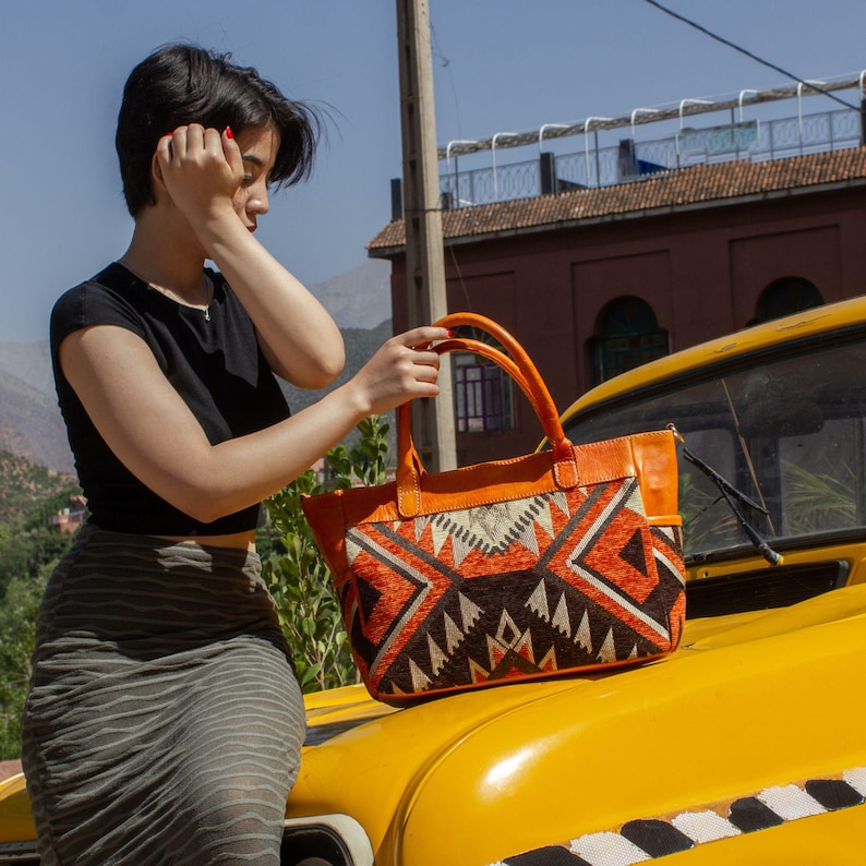 May include: A rectangular handbag featuring orange leather handles and trim, with a central panel displaying a geometric pattern in shades of orange, brown, and cream. The bag has two top handles and a structured base, showcasing a distinct woven texture.