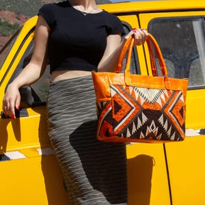 May include: A tote bag featuring orange leather handles and trim, with a central panel displaying a geometric pattern in shades of orange, black, and cream. The bag has two top handles and a structured rectangular shape with side pockets.
