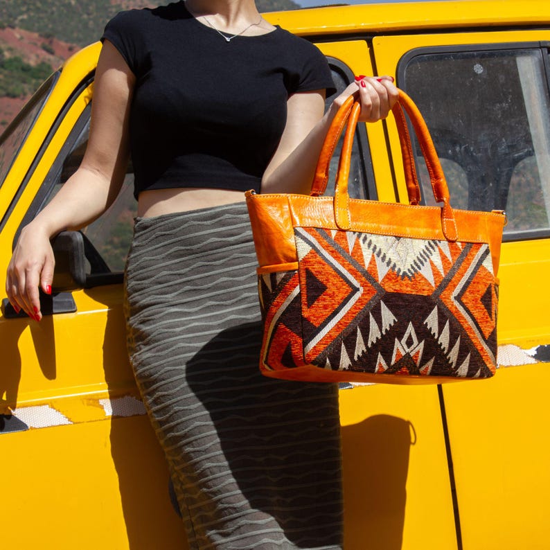 May include: A tote bag featuring orange leather handles and trim, with a central panel displaying a geometric pattern in shades of orange, black, and cream. The bag has two top handles and a structured rectangular shape with side pockets.