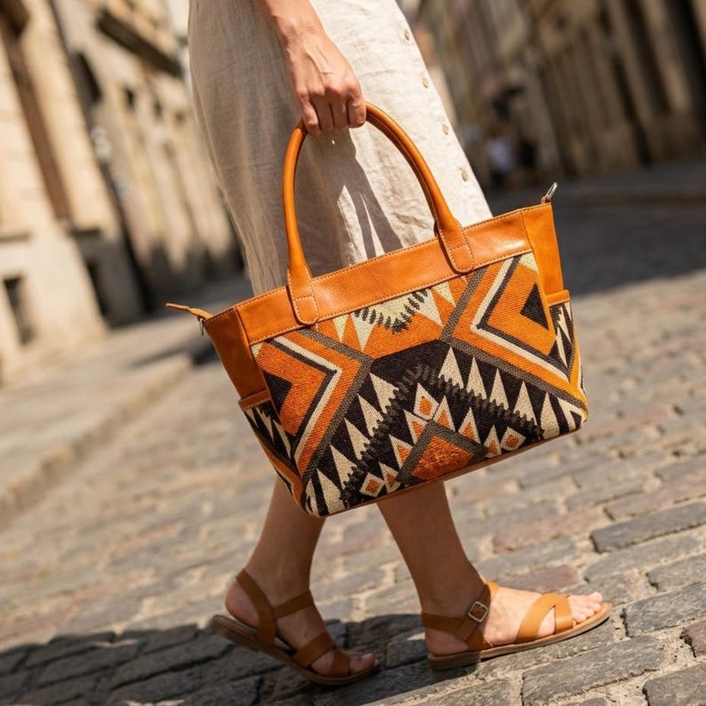 May include: A tote bag featuring tan leather handles and trim, with a central panel displaying a geometric pattern in shades of orange, black, and cream. The bag has a rectangular shape with side pockets and a top zipper closure.