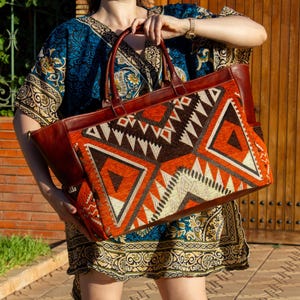 May include: A large rectangular tote bag featuring a vibrant geometric pattern in shades of burnt orange, dark brown, and cream. The bag is constructed with smooth brown leather handles, side panels, and trim, showcasing a structured design.