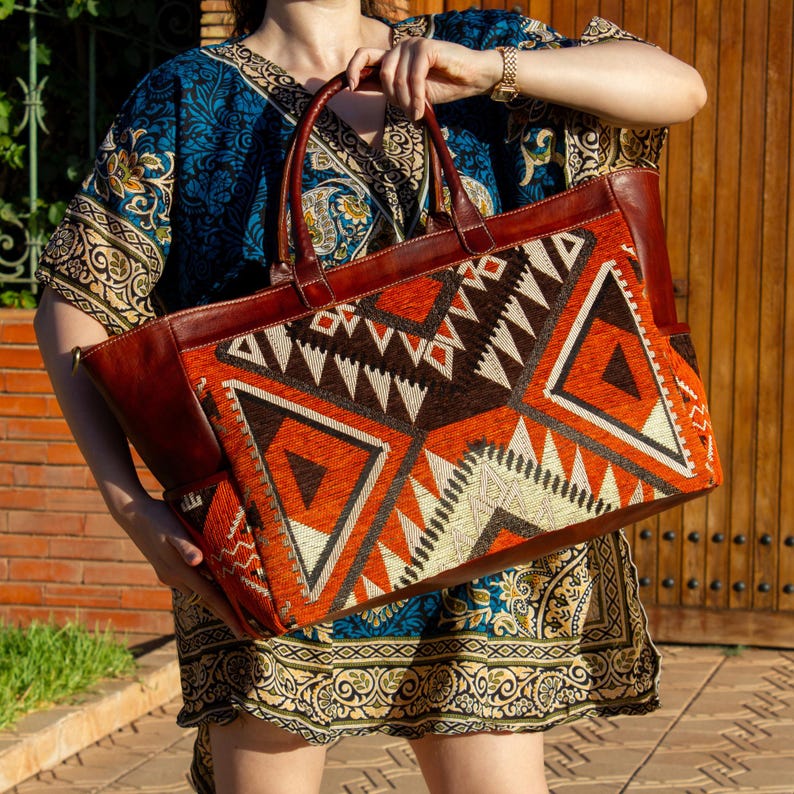 May include: A large rectangular tote bag featuring a vibrant geometric pattern in shades of burnt orange, dark brown, and cream. The bag is constructed with smooth brown leather handles, side panels, and trim, showcasing a structured design.