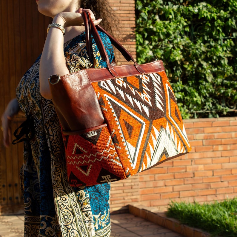 May include: A large tote bag featuring a combination of smooth brown leather and a woven fabric panel with a geometric pattern in shades of orange, brown, and white. The bag includes two leather handles and a side pocket made of brown leather.