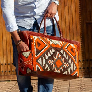 May include: A large leather tote bag featuring a vibrant orange, brown, and cream geometric patterned fabric panel on the front. The bag includes two leather handles, a side pocket, and a smooth brown leather base and trim construction.