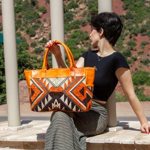 May include: A large tote bag featuring a vibrant geometric pattern in shades of orange, brown, and cream. The bag is constructed with smooth orange leather handles and trim, showcasing a structured rectangular shape with side pockets.