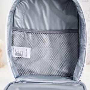 Puede incluir: Vista abierta de una bolsa de almuerzo aislada blanca con interior gris. La bolsa tiene un bolsillo de malla y una etiqueta con texto. El interior est&aacute; forrado con un material aislante reflectante.